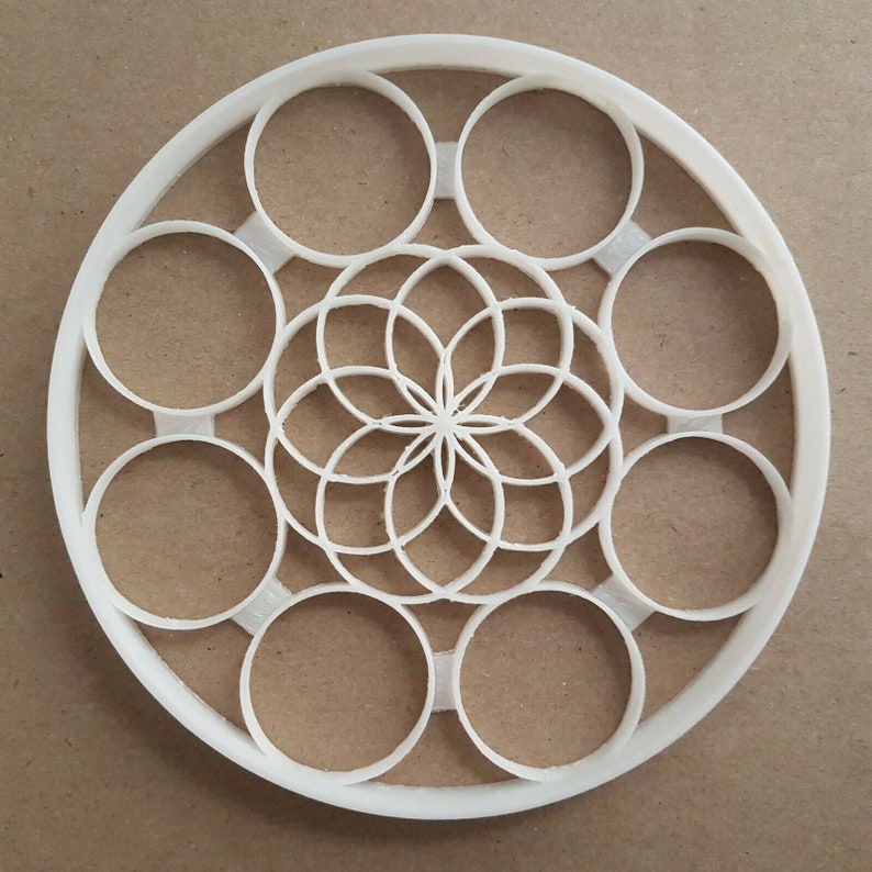 May include: White plastic cookie cutter in the shape of a circle with a geometric design in the center. The design features a flower with multiple layers of petals.