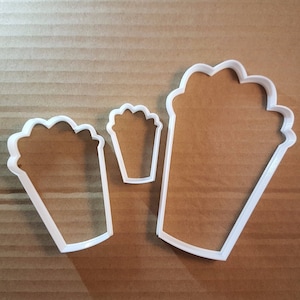 May include: Three white plastic cookie cutters shaped like popcorn containers. The largest cutter is about 5 inches tall and the smallest is about 2 inches tall.