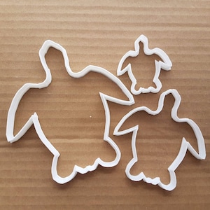May include: Three white plastic turtle cookie cutters of different sizes. The largest turtle is on the left, the smallest turtle is on the right, and the middle-sized turtle is in the centre.