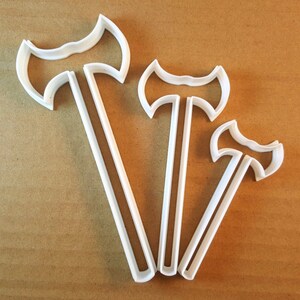May include: Three white plastic cookie cutters shaped like axes of different sizes. The cutters are arranged in a row from largest to smallest.