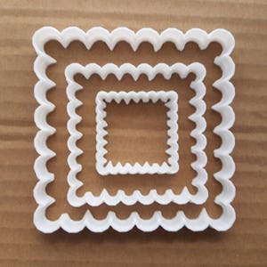 May include: A set of three white plastic cookie cutters shaped like squares with scalloped edges. The cutters are nested inside each other, with the largest cutter on the bottom and the smallest cutter on top.