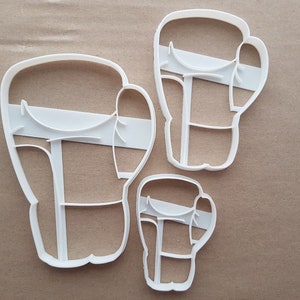 Puede incluir: Tres cortadores de galletas de plástico blanco con forma de guantes de boxeo. El cortador más grande mide aproximadamente 10 cm de alto, el mediano aproximadamente 7,5 cm y el más pequeño aproximadamente 5 cm.