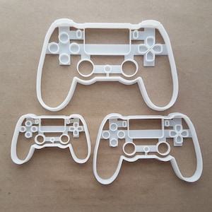 May include: Three white plastic cookie cutters shaped like a video game controller. The largest cutter is approximately 18 cm wide and 10 cm tall. The two smaller cutters are approximately 10 cm wide and 5 cm tall.