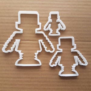 Könnte beinhalten: Vier weiße Ausstecher aus Kunststoff in Form einer blockartigen Person. Die Ausstecher sind auf einer braunen Kartonoberfläche angeordnet. Die Ausstecher sind unterschiedlich groß, der größte ist etwa 10 cm hoch.