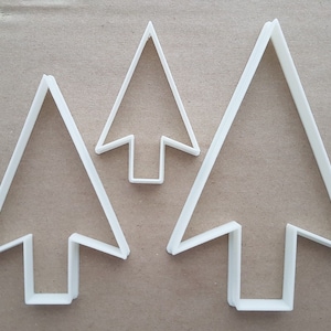 May include: Three white plastic cookie cutters shaped like Christmas trees. The cutters are different sizes.