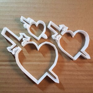 May include: Three white plastic cookie cutters shaped like hearts with a dagger through them.