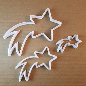 Op de afbeelding: Drie witte plastic koekjesvormen in de vorm van vallende sterren. De grootste vorm heeft een vijfpuntige ster aan het einde van de staart. De andere twee vormen zijn kleiner en hebben een vierpuntige ster aan het einde van de staart.