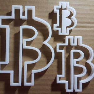 Pode incluir: Dois cortadores de biscoitos de plástico branco em forma de símbolo Bitcoin. Os cortadores estão sobrepostos.