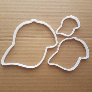 May include: Three white plastic cookie cutters shaped like baseball caps. The largest cutter is about 4 inches wide. The cutters are on a brown surface.