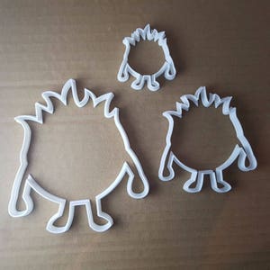 May include: Three white plastic cookie cutters shaped like cartoon monsters with spiky hair and round bodies. The cutters are different sizes.