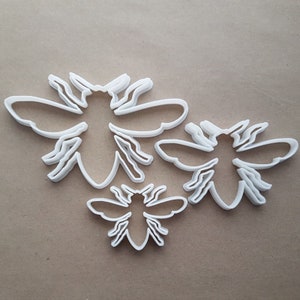May include: Three white plastic bee-shaped cookie cutters of different sizes. The largest cutter is in the center of the image, with the two smaller cutters on either side.