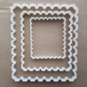 May include: Three white, square cookie cutters with a wavy, scalloped edge. The cutters are nested, each one smaller than the last. They are made of plastic and are arranged on a light brown surface. Ideal for baking.