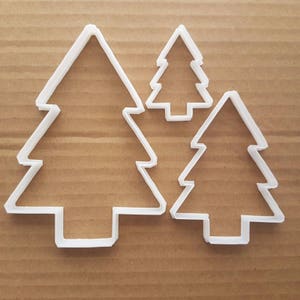 Puede incluir: Tres cortadores de galletas de plástico blanco en forma de árbol de Navidad de diferentes tamaños. El cortador más grande está a la izquierda, el más pequeño a la derecha y el cortador de tamaño mediano en el centro.