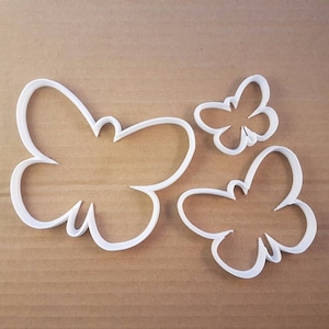 May include: Three white butterfly-shaped cookie cutters of varying sizes. The cutters are made of a solid white material and are arranged on a brown surface. The largest cutter is approximately 4 inches wide.