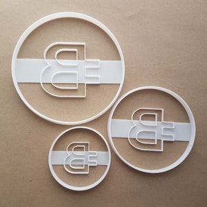 May include: Three white plastic cookie cutters shaped like a Bitcoin symbol. The cutters are different sizes.