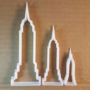 Puede incluir: Tres cortadores de galletas de plástico blanco con forma de rascacielos. Los cortadores son de diferentes tamaños, siendo el más grande el más alto.