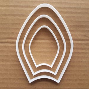 Może przedstawiać: Zestaw trzech białych plastikowych foremek do ciasteczek w kształcie zaokrąglonego, spiczastego owalu. Formy są włożone jedna w drugą, przy czym największa forma jest na zewnątrz, a najmniejsza wewnątrz.