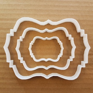 以下が含まれることがあります： 装飾的なフレームの形をした白いプラスチック製のクッキーカッターが3つあります。カッターは互いにネストされており、最も小さいカッターが中央にあります。