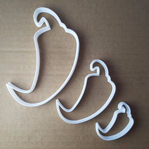 May include: Three white plastic chili pepper-shaped cookie cutters in varying sizes. The cutters are arranged diagonally, with the largest at the top left and the smallest at the bottom right. The background is a solid, neutral brown.