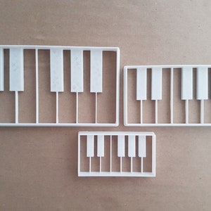 Puede incluir: Tres cortadores de galletas de plástico blanco con forma de teclado de piano. Los cortadores son de diferentes tamaños, el más grande tiene 7 teclas, el mediano 5 teclas y el más pequeño 4 teclas.