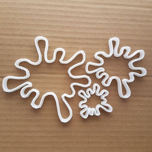 May include: Three white plastic cookie cutters shaped like stylized abstract flowers. The largest cutter is in the center and the two smaller cutters are on either side.