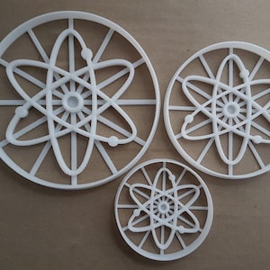 May include: Three white plastic cookie cutters shaped like an atom. The cutters are different sizes, with the largest cutter being about 6 inches in diameter.