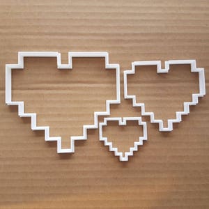 Könnte beinhalten: Drei weiße Plastik-Ausstecher in Form von pixelartigen Herzen. Das größte Herz befindet sich oben, das mittelgroße Herz rechts und das kleinste Herz unten.