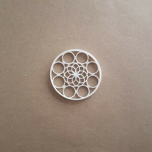 May include: White plastic cookie cutter in the shape of a circle with a geometric design in the center. The design features a flower with multiple layers of petals.