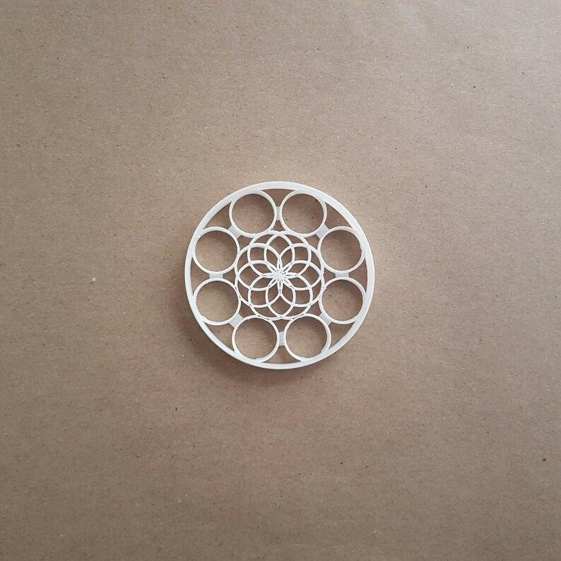 May include: White plastic cookie cutter in the shape of a circle with a geometric design in the center. The design features a flower with multiple layers of petals.