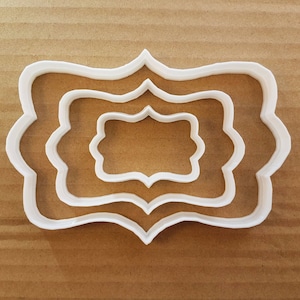 Puede incluir: Tres cortadores de galletas de plástico blanco en forma de rectángulo festoneado. Los cortadores están anidados uno dentro del otro, con el cortador más grande en la parte inferior y el más pequeño en la parte superior.