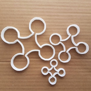 Puede incluir: Un juego de cortadores de galletas de plástico blanco con forma de círculos conectados. Los cortadores están dispuestos en un patrón sobre una superficie marrón.