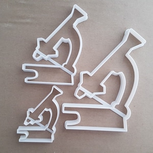 May include: Three white plastic cookie cutters shaped like microscopes. The cutters are layered on top of each other.