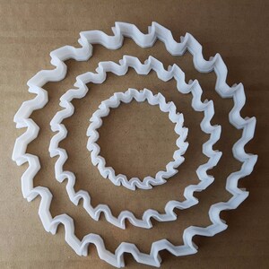 May include: Three white plastic cookie cutters in the shape of a scalloped circle. The cutters are nested inside each other, with the largest cutter on the outside and the smallest cutter on the inside.