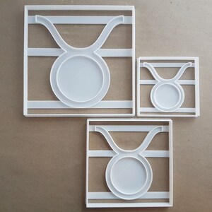 May include: Three white plastic cookie cutters shaped like the astrological sign Taurus. The cutters are different sizes, with the largest cutter at the top of the image and the smallest cutter at the bottom.