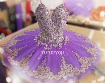 May include: A purple and gold ballet tutu with a lace trim and a bodice featuring a floral pattern. The tutu is made of tulle and has a full skirt. The bodice is fitted and has a sweetheart neckline. The tutu is decorated with sequins and beads. The text "TUTU2YOU" is visible on the skirt.