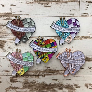 May include: Five enamel pins shaped like hearts with a yarn ball and knitting needles inside. Each pin has a white banner with the text "PROCRASTINATOR" on it. The hearts are different colors and patterns: red and white checkered, purple and blue striped, rainbow, green and pink checkered, and white with a light blue and pink pattern.