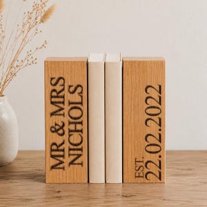 May include: Two rectangular light brown wooden bookends with dark engraved text. The left bookend reads MR & MRS NICHOLS and the right bookend reads EST. 22.02.2022. Two plain cream-colored books are positioned vertically between the wooden blocks.