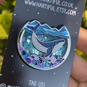 May include: A round enamel pin featuring a blue whale swimming in the ocean. The pin has a silver border and is set against a dark background with the text "WWW.HARTIFUL.CO.UK" and "WWW.HARTIFUL.ETSY.COM". The pin is held by a hand.