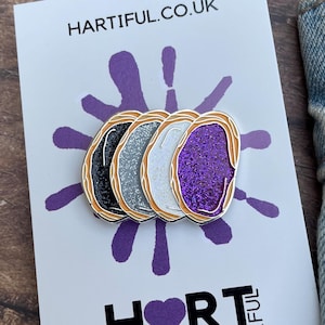 May include: A set of four enamel pins in black, silver, white, and purple glitter designs, each with a gold border. The pins are displayed on a white card with the text "HARTIFUL.CO.UK" and a purple splatter design. The word "HORT" is also visible.