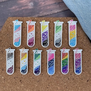 Puede incluir: Una colección de once pines de tubos de ensayo coloridos. Cada pin presenta un diseño único con una variedad de colores, incluyendo azul, naranja, morado, verde y amarillo. Los pines están colocados sobre un fondo de corcho marrón.