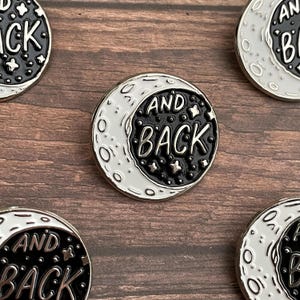 May include: A black and white enamel pin with a crescent moon and stars. The text "AND BACK" is written in a white outline on the black moon.