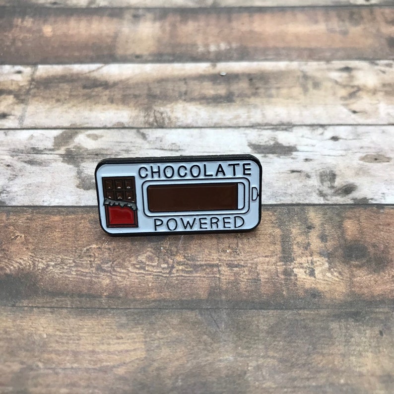 K&ouml;nnte beinhalten: Eine wei&szlig;e Emaille-Anstecknadel mit einer Schokoladentafel-Grafik und dem Text "Chocolate Powered". Die Schokoladentafel ist teilweise gegessen und die Batterieanzeige ist voll.