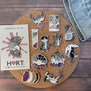 May include: A collection of enamel pins with various designs, including a phoenix, cats, a dachshund, crystals, and a tarot card. The pins feature a color palette of black, white, purple, and silver. A business card with the text "HARTIFUL.CO.UK" is also present.