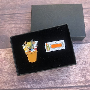 Könnte beinhalten: Eine schwarze Box mit weißem Hintergrund, die zwei Emaille-Pins enthält. Ein Pin ist ein Eimer gefüllt mit Pinseln, Farbtuben und Bleistiften. Der andere Pin ist ein Batteriesymbol, auf dem "Art Powered" steht.