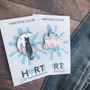 May include: Two enamel pins on white cards with a blue splatter design. The first pin features a black cat sitting on a crescent moon with pink and blue crystals. The second pin features a pink and blue crystal cluster. The cards have the text "HARTIFUL.CO.UK" and "HRTIFUL" with social media handles.