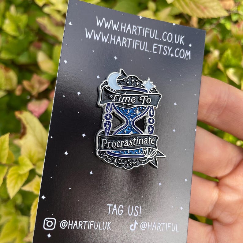 May include: A black enamel pin with a blue hourglass design, the text "Time To Procrastinate" is displayed on a banner. The pin also features a crescent moon and stars. The pin is on a black card with the website address www.hartiful.co.uk and www.hartiful.etsy.com.