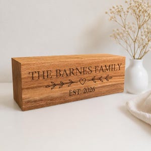 May include: A rectangular light brown wooden block with a natural grain finish. The front face is engraved with the text THE BARNES FAMILY, a central heart icon flanked by leafy branches, and the text EST. 2026 in a dark, clear sans-serif font.