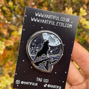 May include: A silver-colored pin featuring a black wolf howling at a full moon, set against a starry night sky. The pin is on a black card with the website address and social media handles.