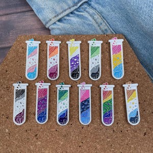 May include: A collection of eleven colorful test tube pins. Each pin features a different design with a variety of colors, including pink, blue, yellow, green, and purple. The pins have a white outline and are arranged on a brown surface.