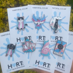 May include: A collection of enamel pins on backing cards. The pins feature various designs, including a cat, a phoenix, a dachshund, a crystal, a cat on a crescent moon, and a tarot card. The pins have a color scheme of pink, blue, and white. The cards have the text "HARTIFUL.CO.UK".